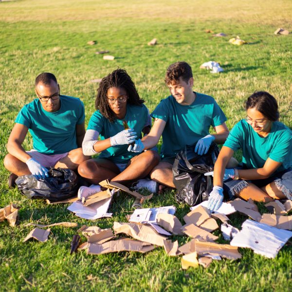 Eco volunteers sorting glass and paper waste on city lawn. Men and woman sitting on grass, picking up litter into plastic bags. Recycling concept