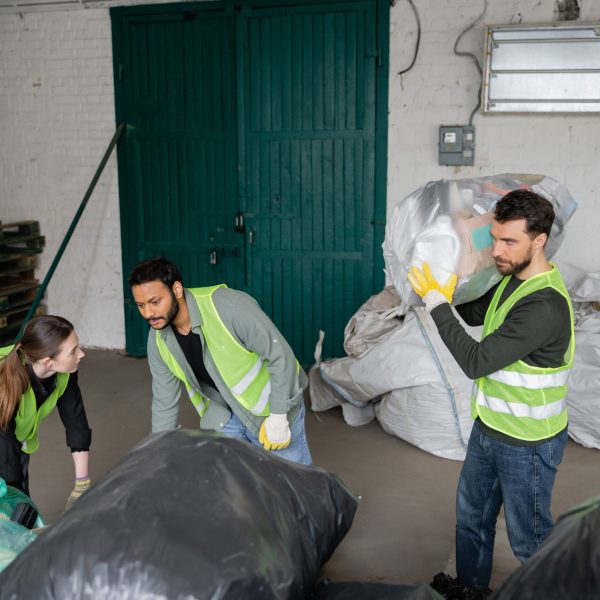 Multiethnic workers in high visibility vests and gloves working with plastic bags near colleague in waste disposal station, garbage sorting and recycling concept